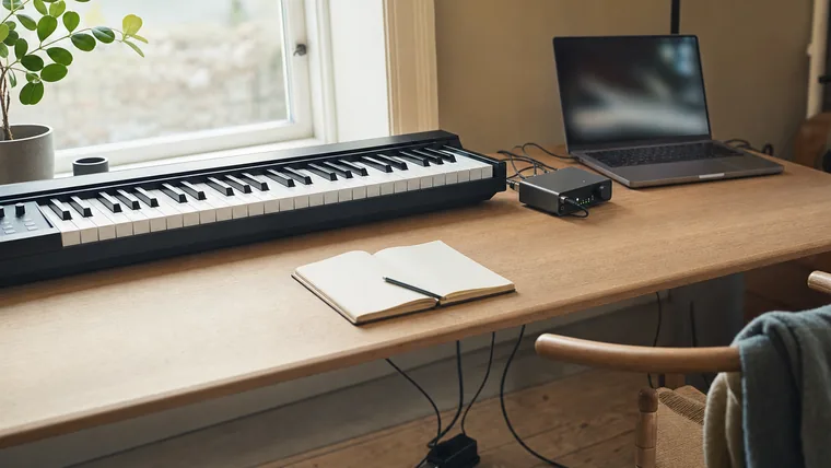 Ett digitalpiano med anslutna MIDI- och pedalkablar på ett skrivbord i ett hemmastudio-hörn.