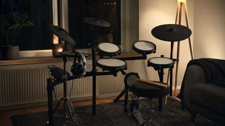 Elektroniskt trumset med nätpads i ett vardagsrumshörn, hörlurar hänger över hi-hat-stativet.